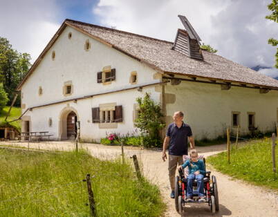 Eine Person nutzt einen geländegängigen 4x4‑Rollstuhl auf einem Naturweg im Freilichtmuseum Ballenberg. Das historische Bauernhaus im Hintergrund zeigt, wie auch unebene Wege dank mietbarem Spezialrollstuhl zugänglich werden.