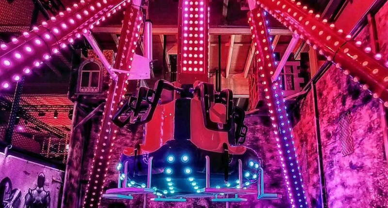 Eine leuchtende Fahrattraktion in einer Indoor-Halle, umgeben von intensiven pinken und violetten Lichteffekten. Die kräftigen LED-Streifen und die futuristische Inszenierung verleihen der Szene eine dynamische, energiegeladene Atmosphäre.